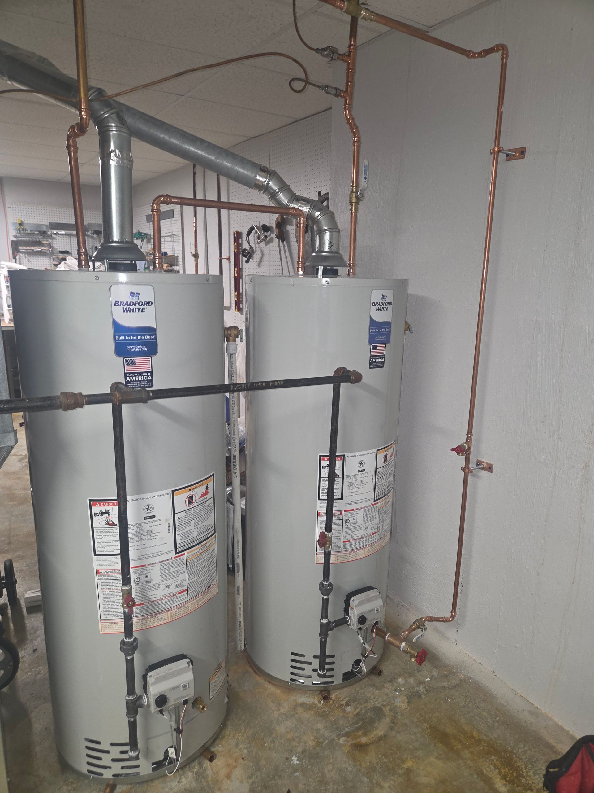 Two water heaters with copper pipes and venting in a utility room, with a concrete floor and white walls.