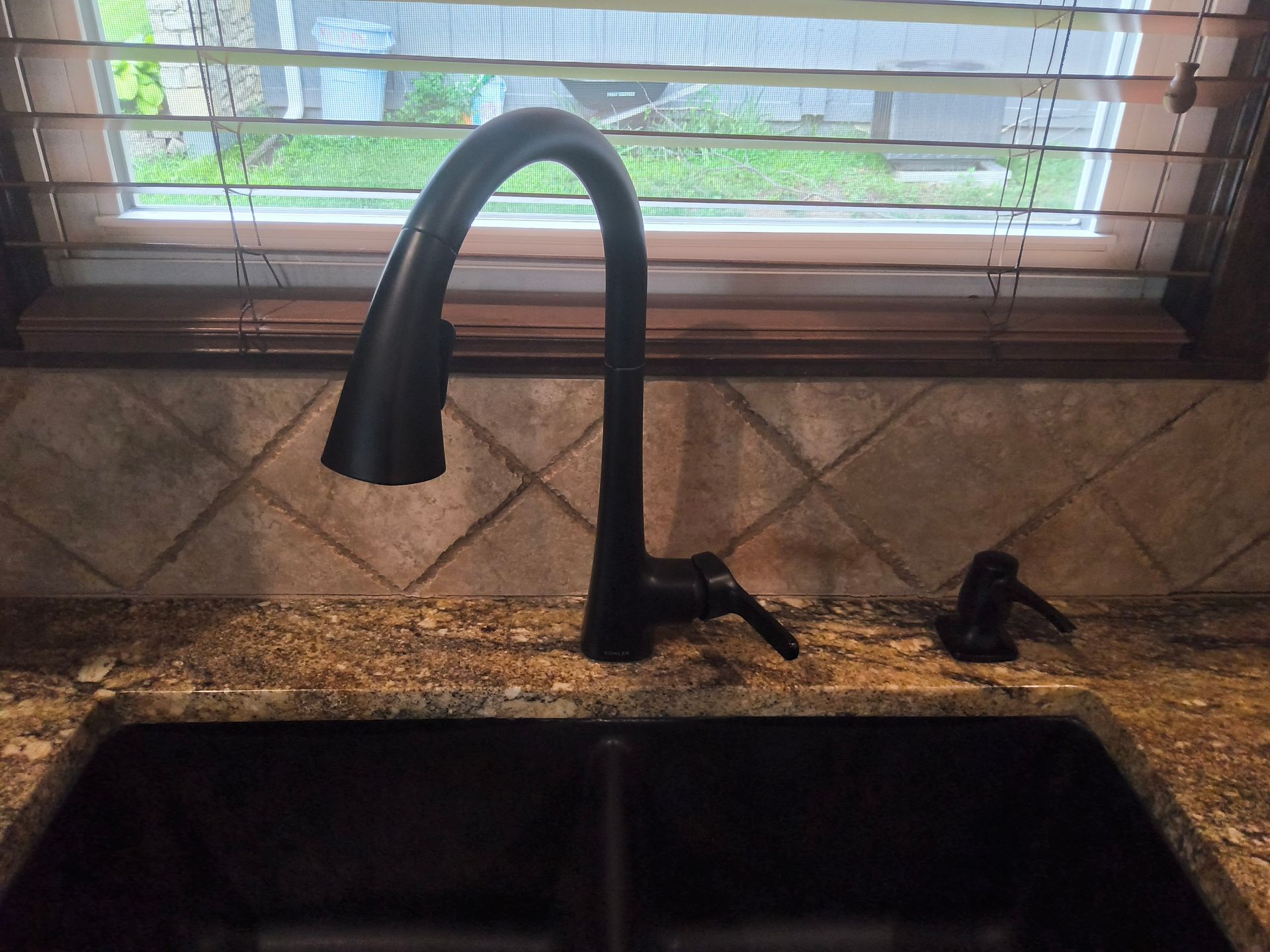 Black kitchen sink with tall matte faucet and window blinds above granite countertop