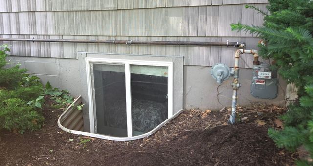 Basement window well with white-framed sliding window under gray shingled siding and utility meters nearby