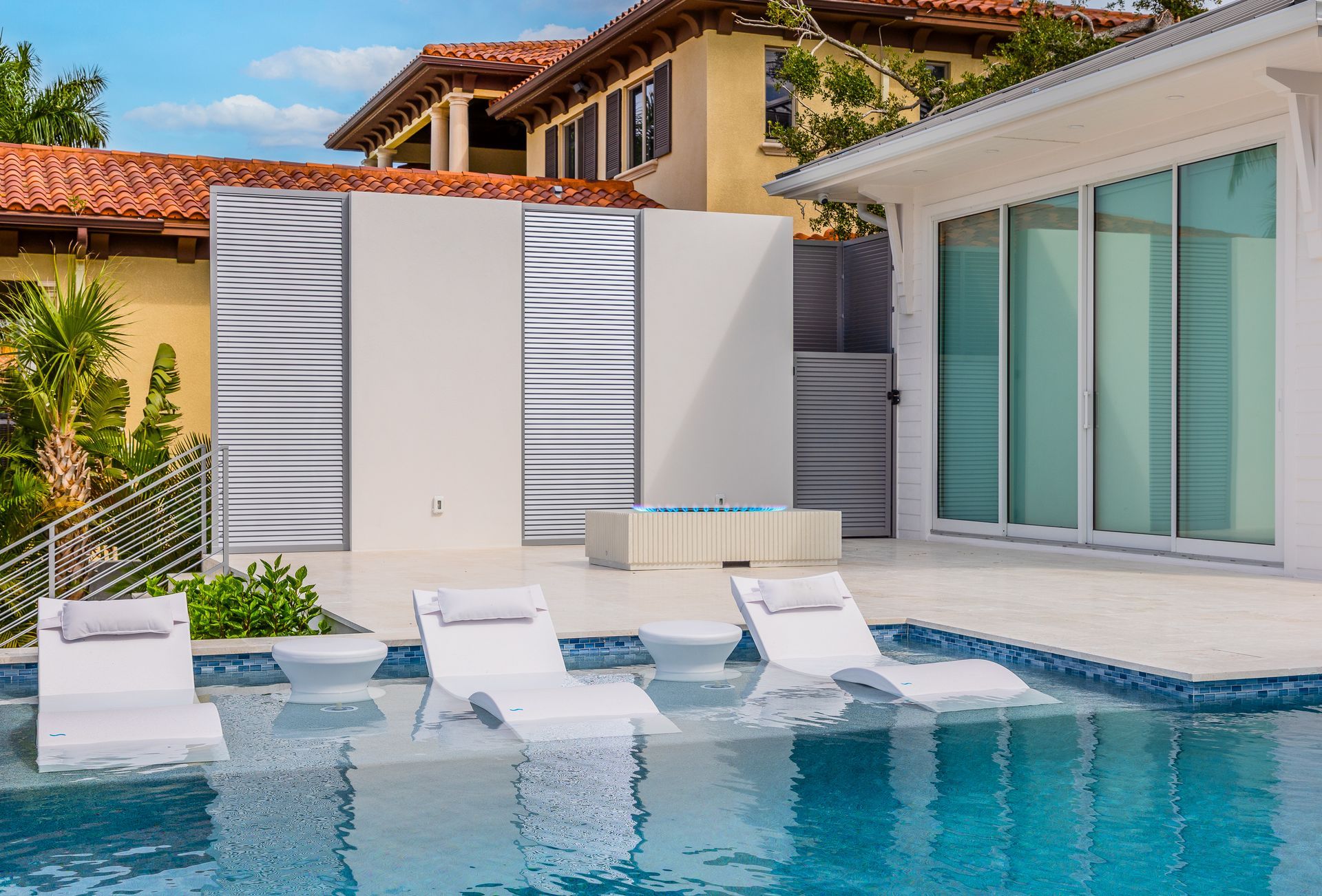 Modern poolside patio with white lounge chairs, blue water, and sliding glass doors