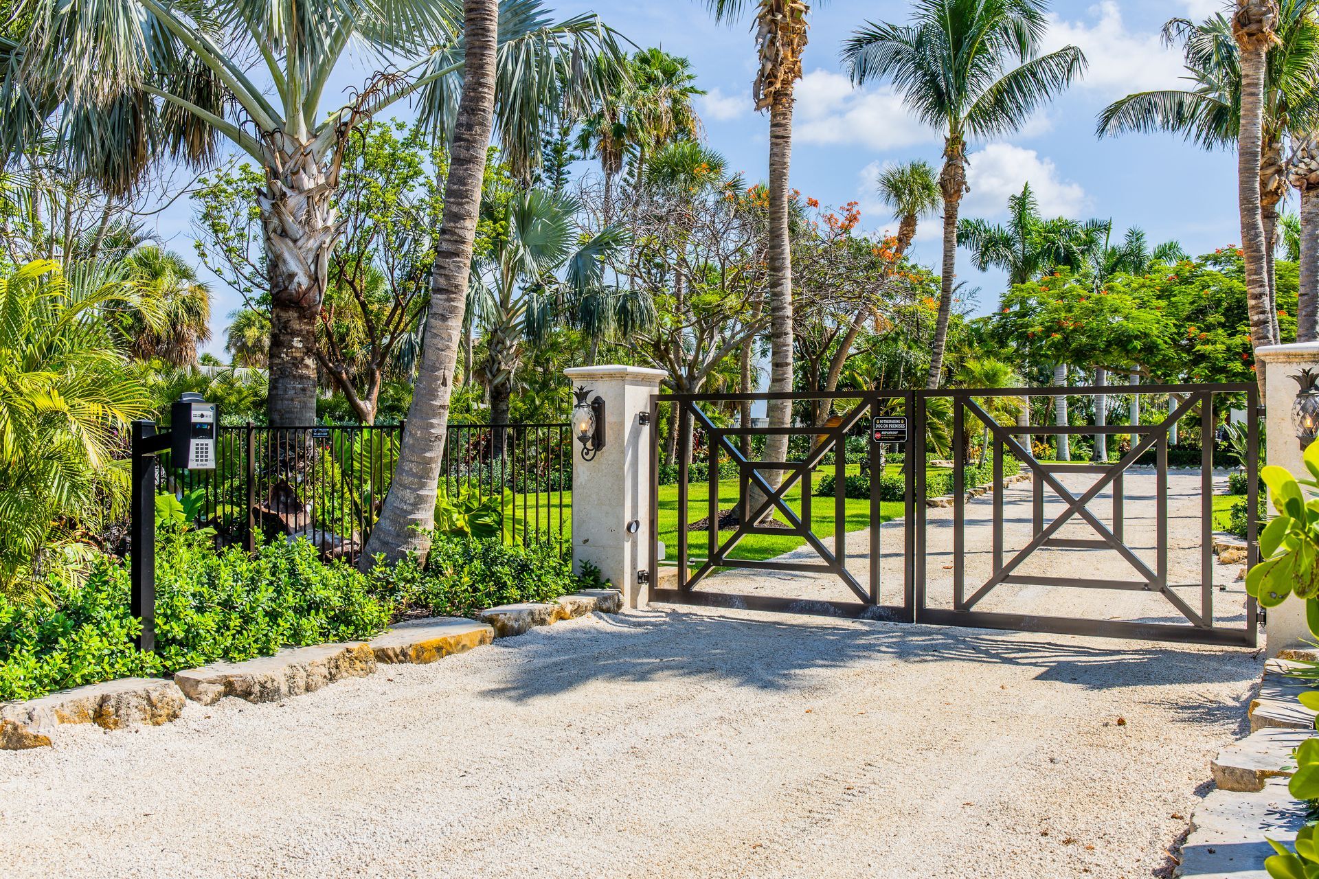 Gravel driveway with black metal gate, stone pillars, and lush tropical trees under a bright sky