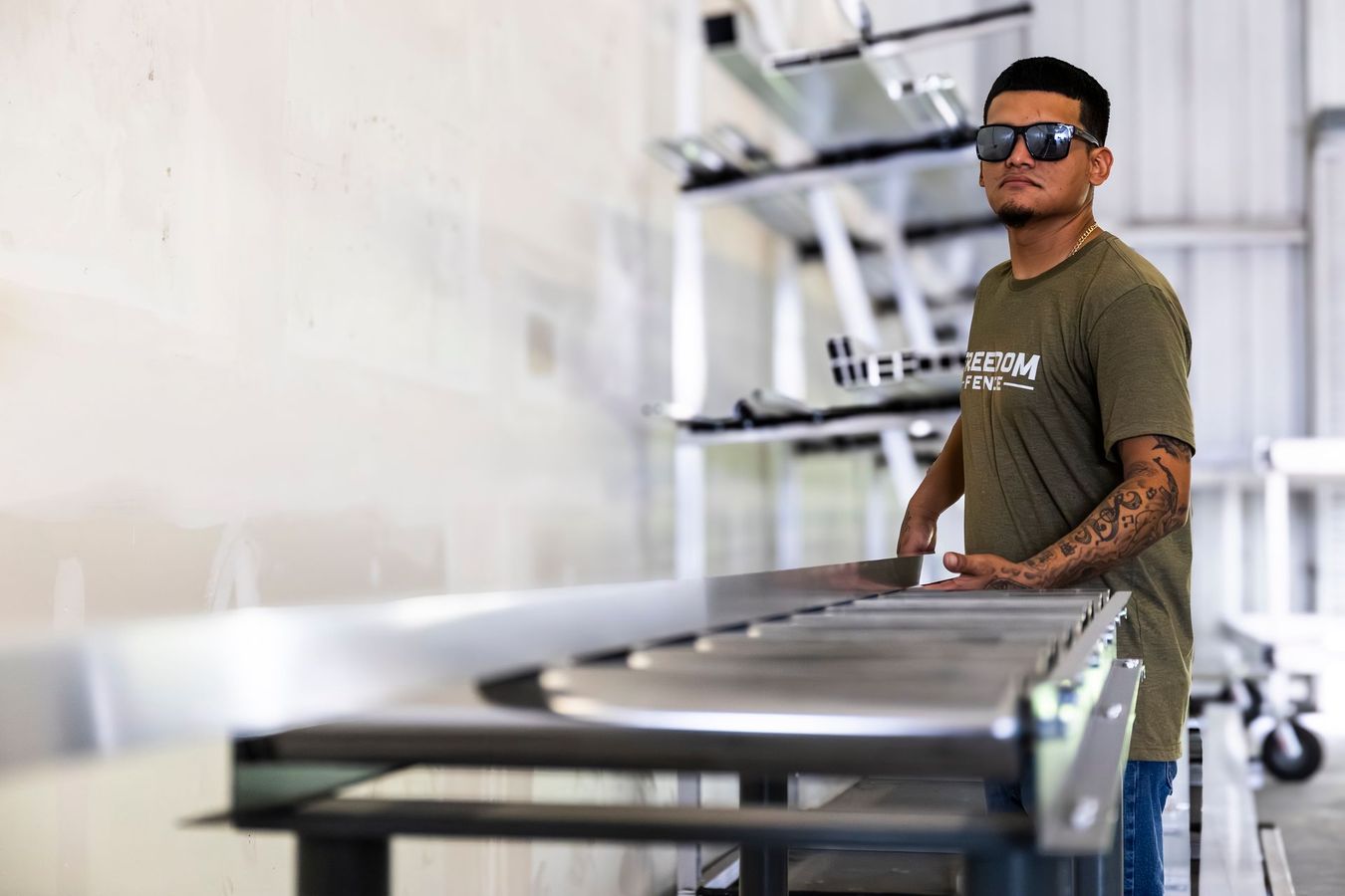 Man standing beside a metal worktable in an industrial workshop, wearing sunglasses and a green shirt.