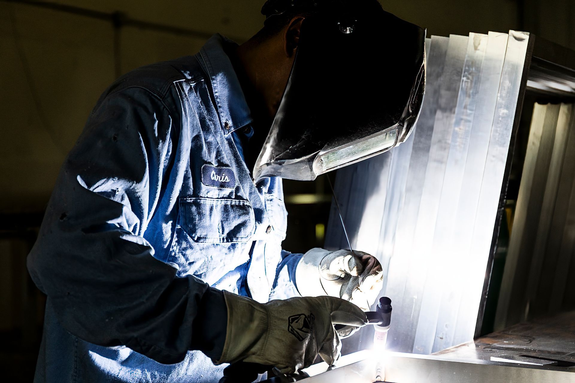 Welder in blue shirt and helmet working on a bright metal piece indoors