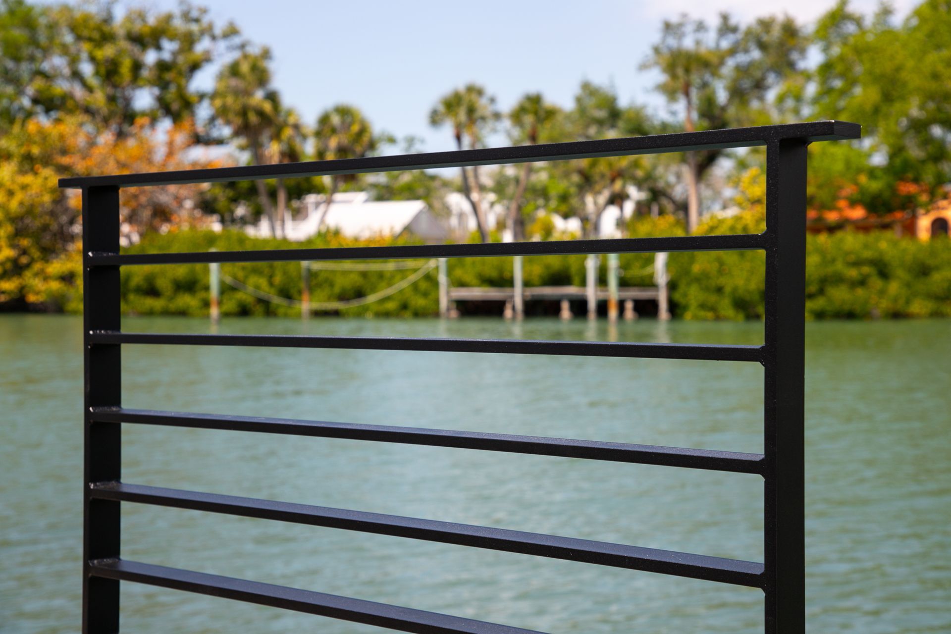 Black metal railing beside a calm lake with trees and a pavilion in the background