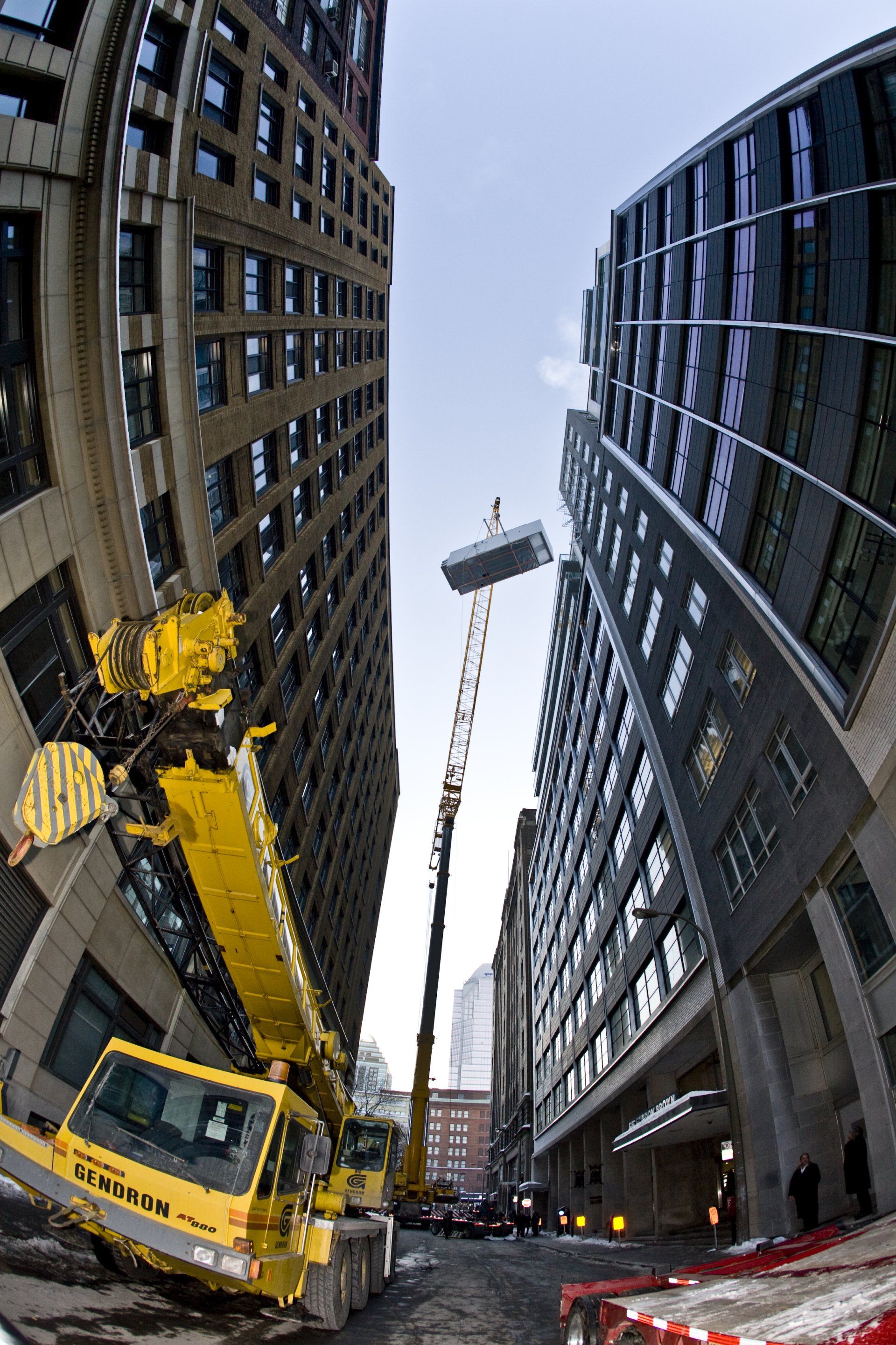 Une grue soulève un objet volumineux entre de hauts immeubles dans une rue de la ville. La grue est jaune.