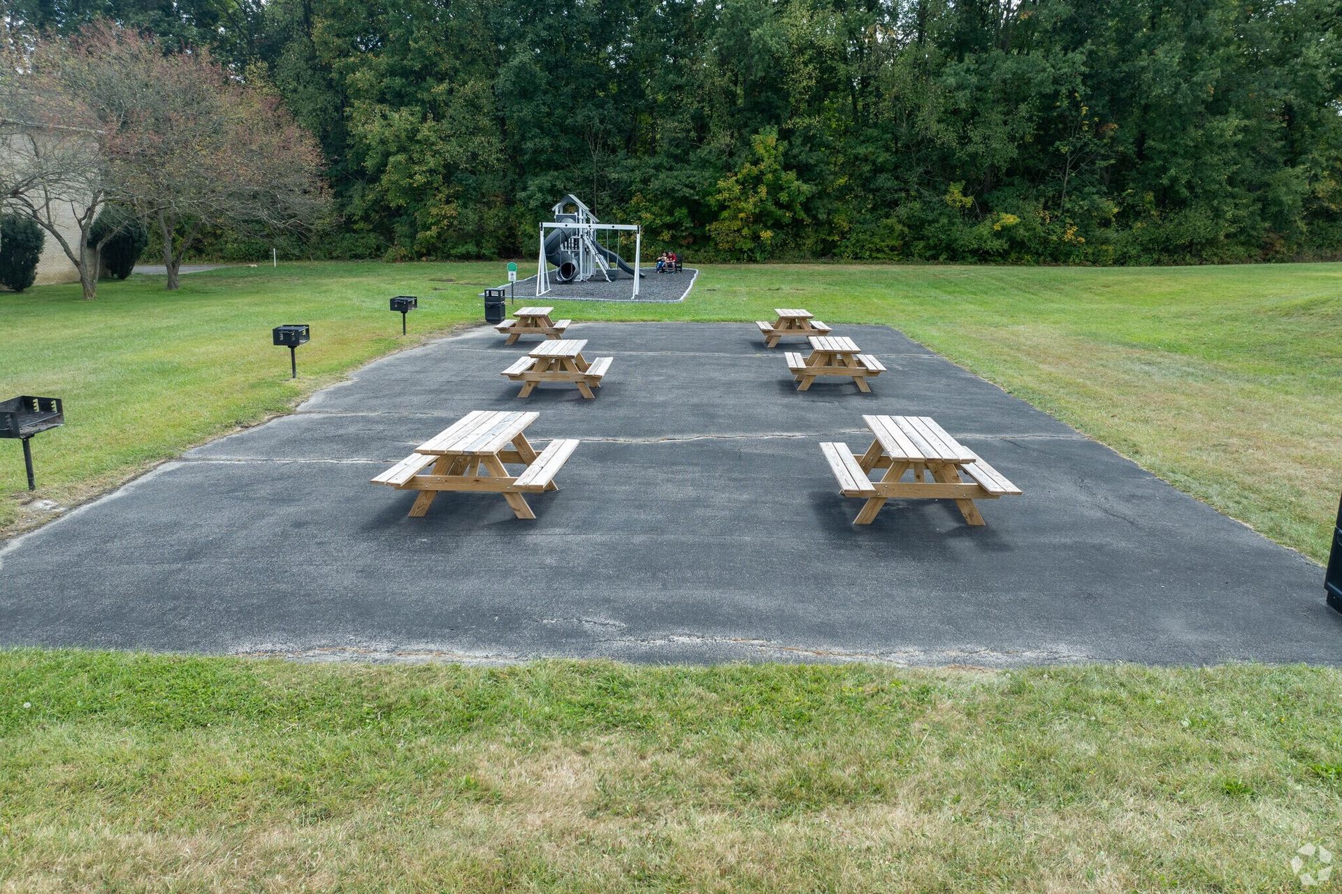 Picnic area with wooden tables on a gravel surface. Grassy surroundings and playground in background.