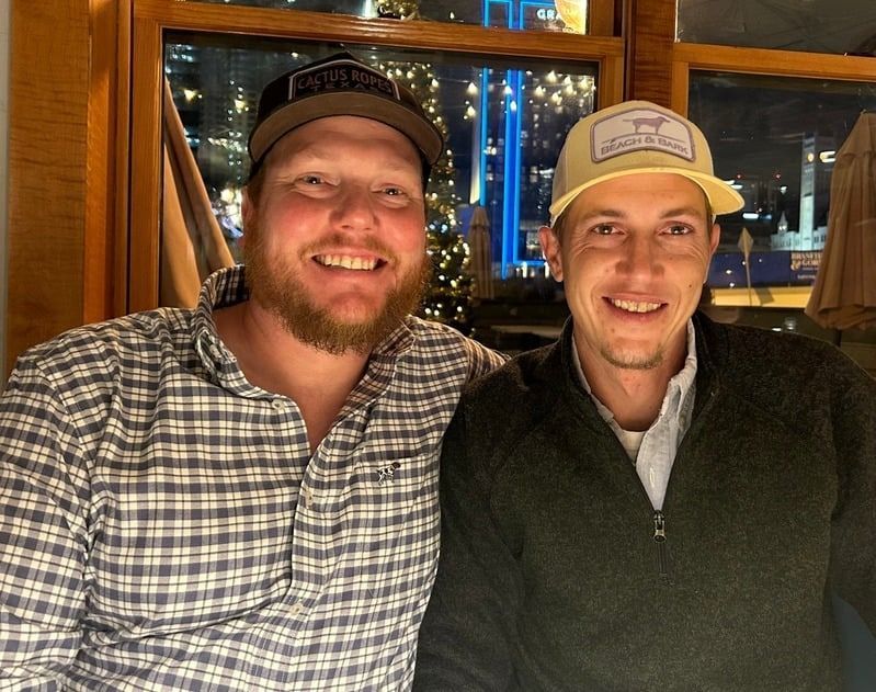 Two smiling people in hats seated indoors by a window at a warmly lit restaurant