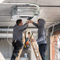 Two workers install a ceiling-mounted HVAC unit on a ladder inside a room under construction.