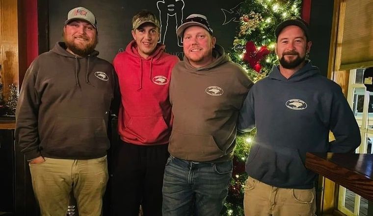 Four people standing together outdoors at night, wearing hoodies and smiling in festive lighting.