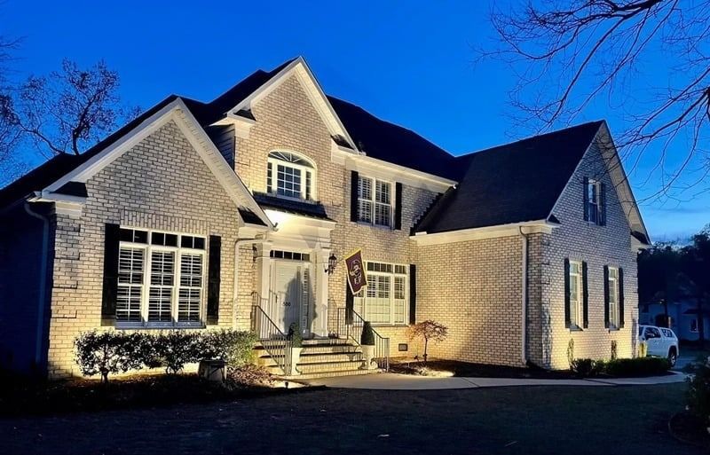 Well-lit brick house at dusk with lit windows and a dark roof under a blue sky