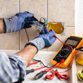 Electrician wiring an outlet with gloves; multimeter and tools on a tiled wall.