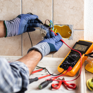 Electrician wiring an outlet with gloves; multimeter and tools on a tiled wall.