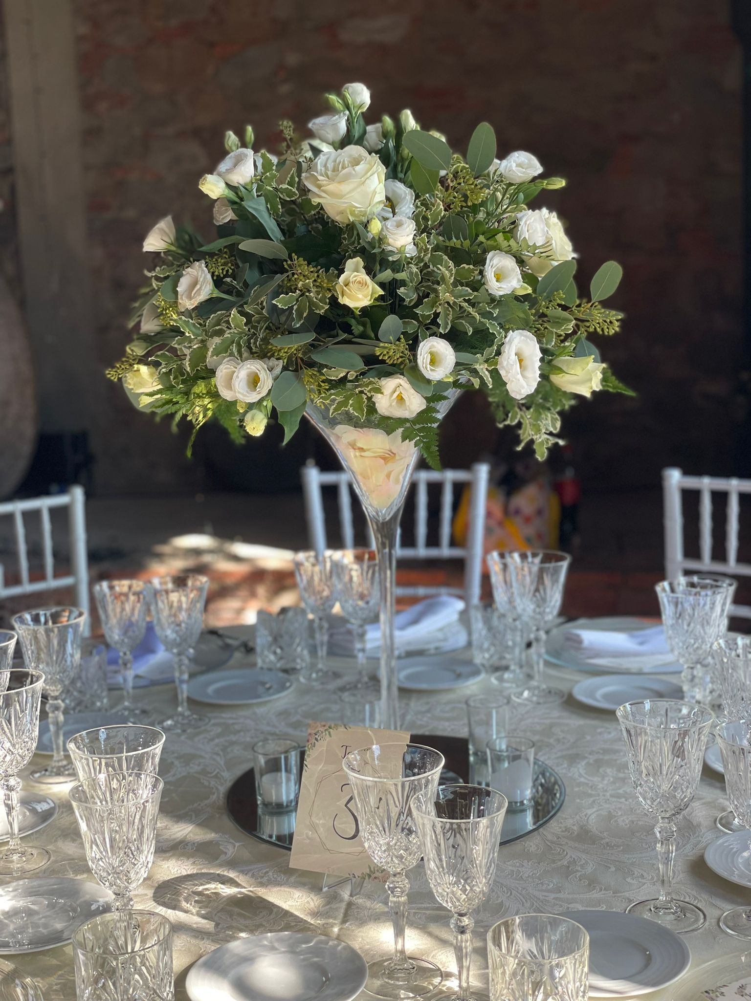 Un tavolo apparecchiato per un ricevimento di nozze con un vaso da martini pieno di fiori bianchi.