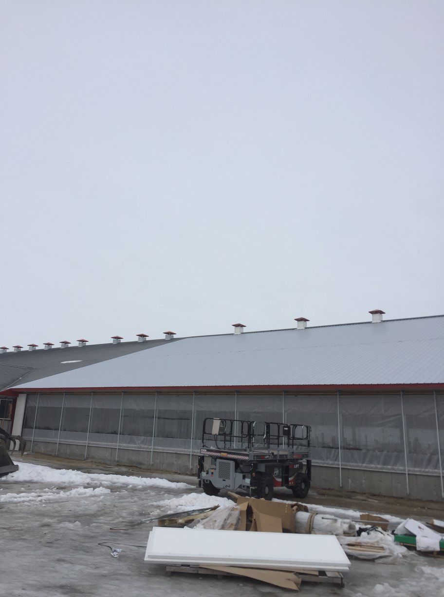 Un bâtiment gris avec un long toit métallique en pente, de multiples ouvertures de ventilation et une nacelle élévatrice garée à l'extérieur sur un sol enneigé et verglacé.