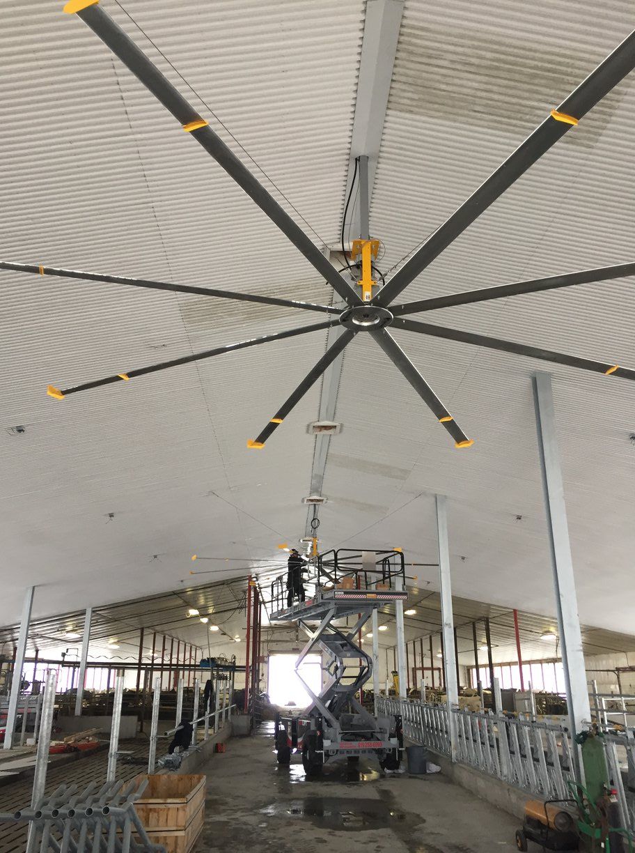 Un ouvrier, sur une nacelle élévatrice, installe un grand ventilateur de plafond industriel à six pales à l'intérieur d'une grange.