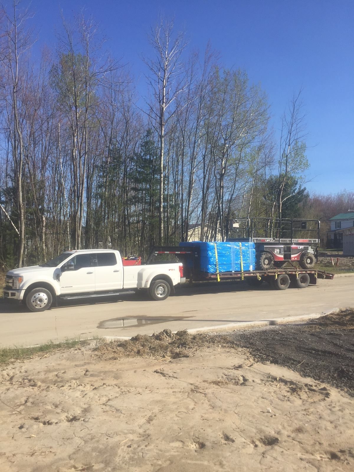 Une camionnette blanche tire une remorque chargée de matériaux emballés en bleu et d'un équipement sur un chemin de terre.