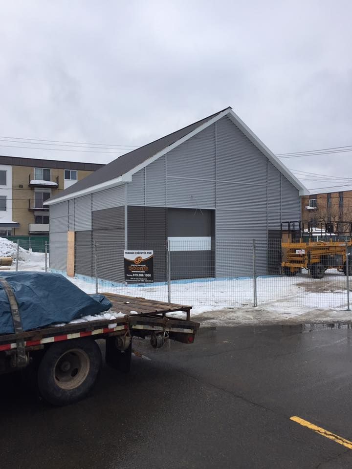 Un bâtiment gris, récemment construit, se dresse sur un terrain enneigé près d'une route, avec des engins de chantier visibles sur le côté.