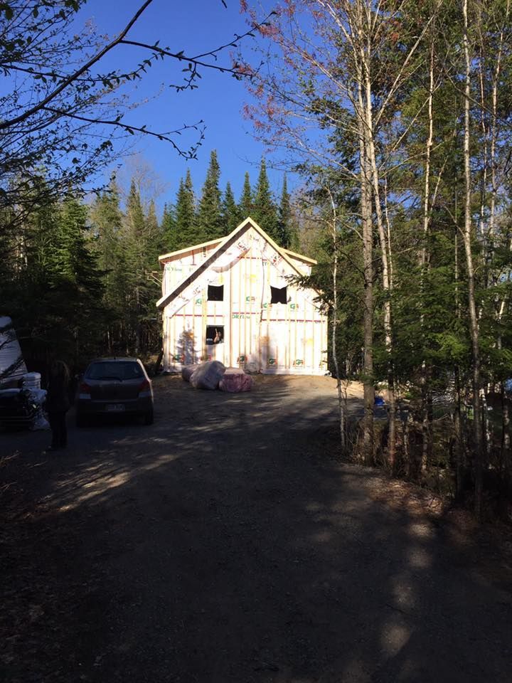 Une maison à deux étages en construction se dresse dans une zone boisée, encadrée par de grands arbres, avec une allée au premier plan.