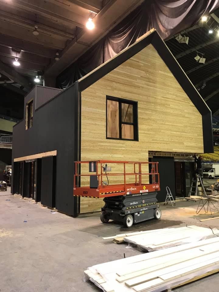 Une maison en construction, avec un bardage en bois et noir, se dresse dans un grand espace intérieur, où l'on aperçoit une nacelle élévatrice rouge.