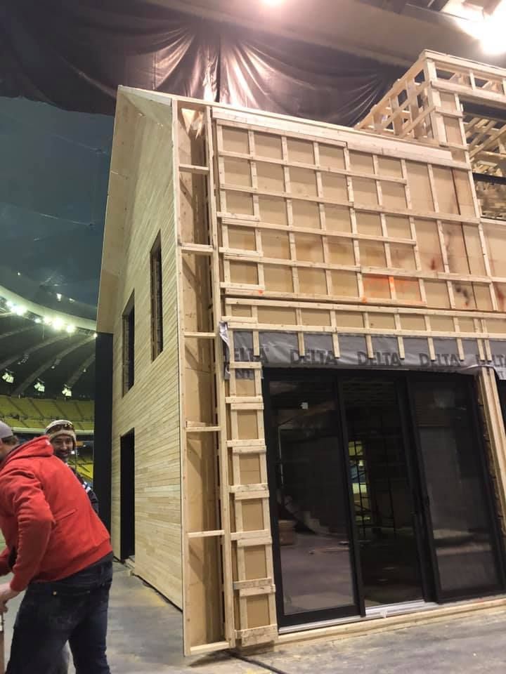 Une personne vêtue d'une veste rouge se tient à côté de la charpente en bois d'une maison en construction à l'intérieur d'un grand stade couvert.