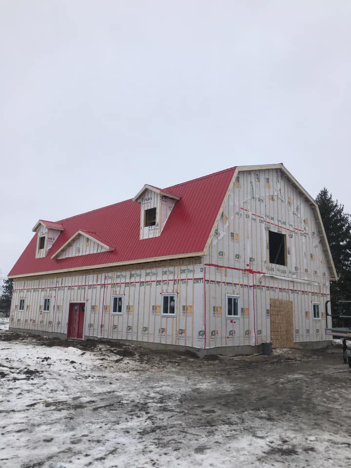 Une grange en construction, avec un revêtement extérieur blanc, un toit en métal rouge et des lucarnes, sur un terrain enneigé.