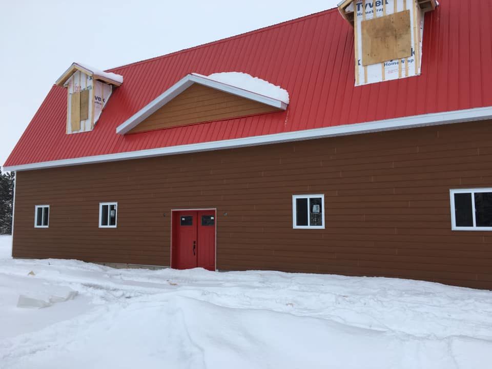 Un bâtiment brun avec un toit en métal rouge et des doubles portes rouges, entouré de neige sous un ciel nuageux.