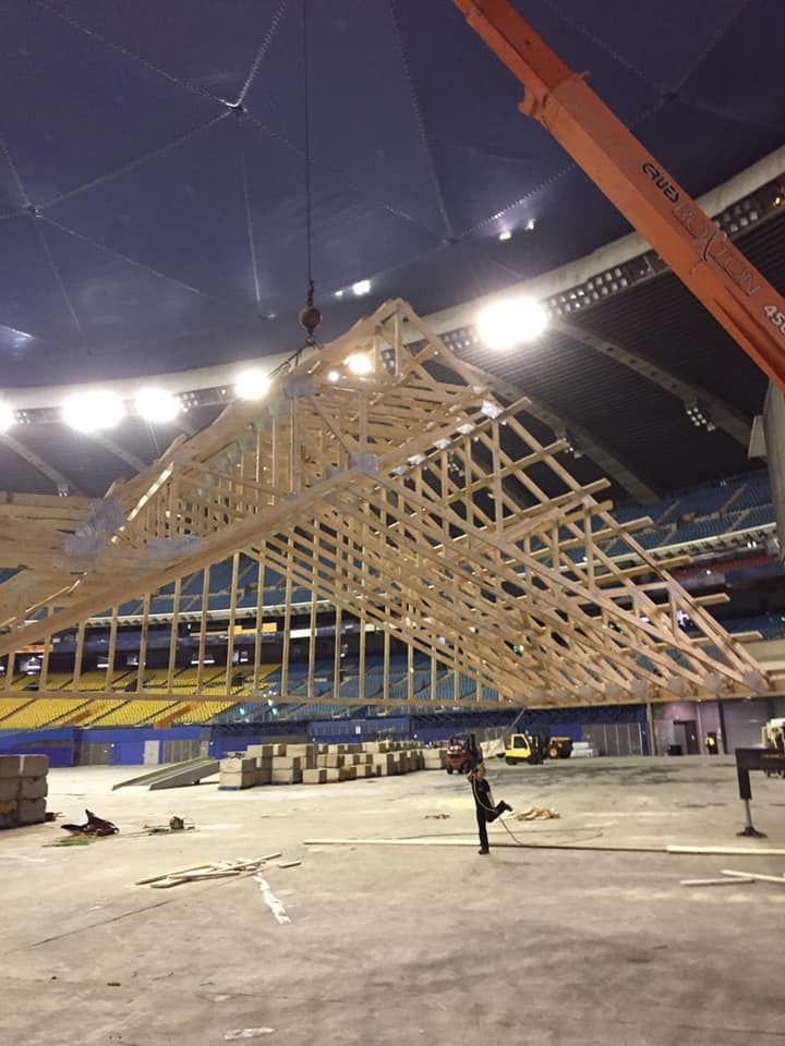 Une grande charpente de toit en bois est suspendue par une grue à l'intérieur d'un stade couvert vide et illuminé.