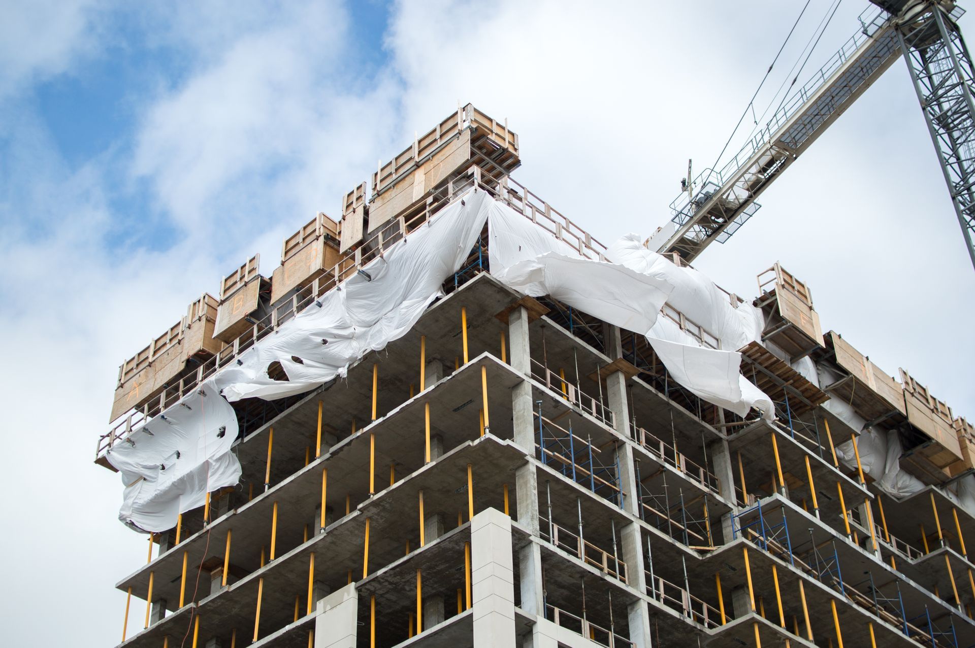 Immeuble de grande hauteur en construction, avec sols en béton, poutres de soutien jaunes et bâche de protection blanche, sous un ciel bleu.