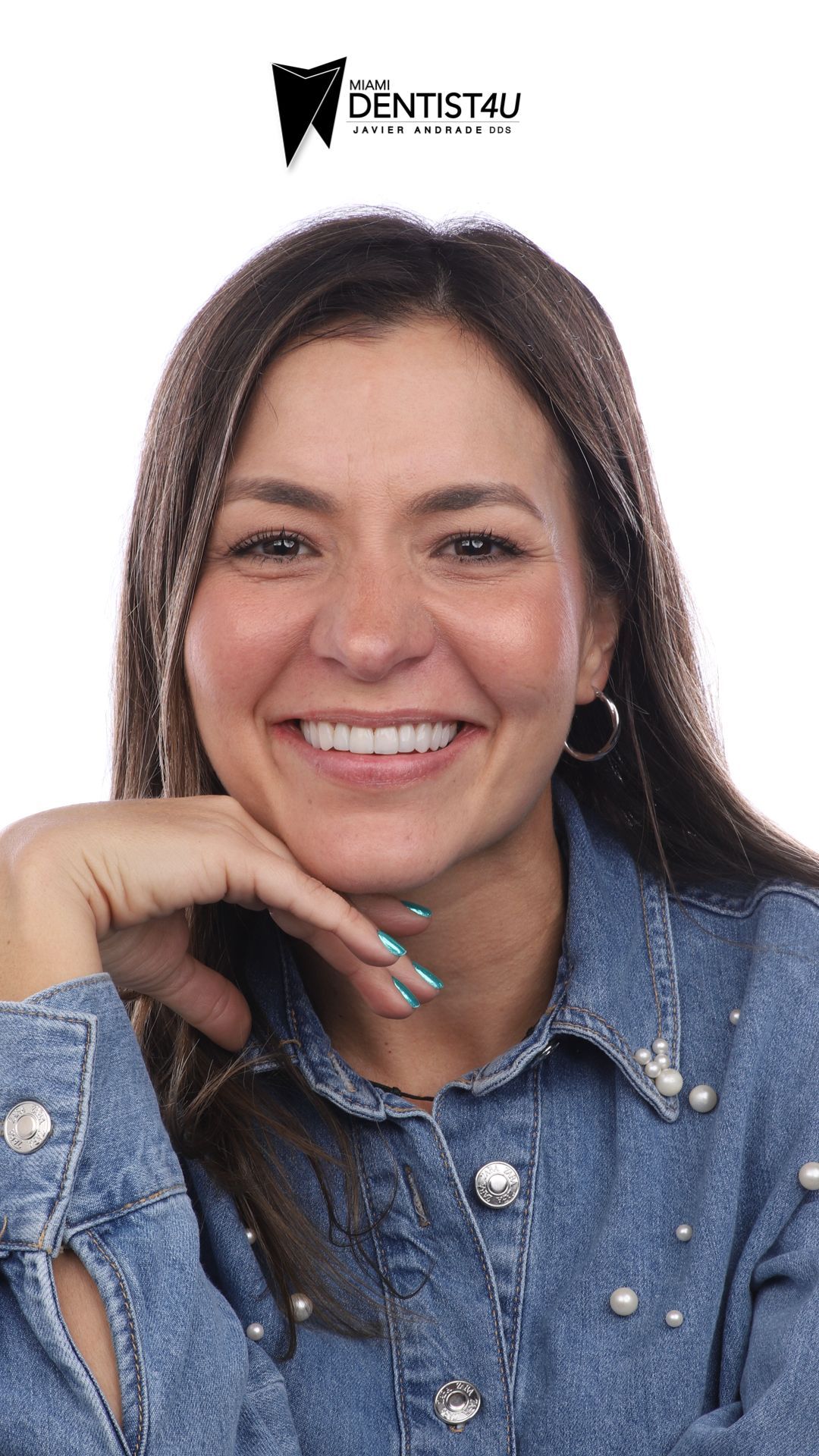 A woman in a denim shirt is smiling with her hand on her chin.