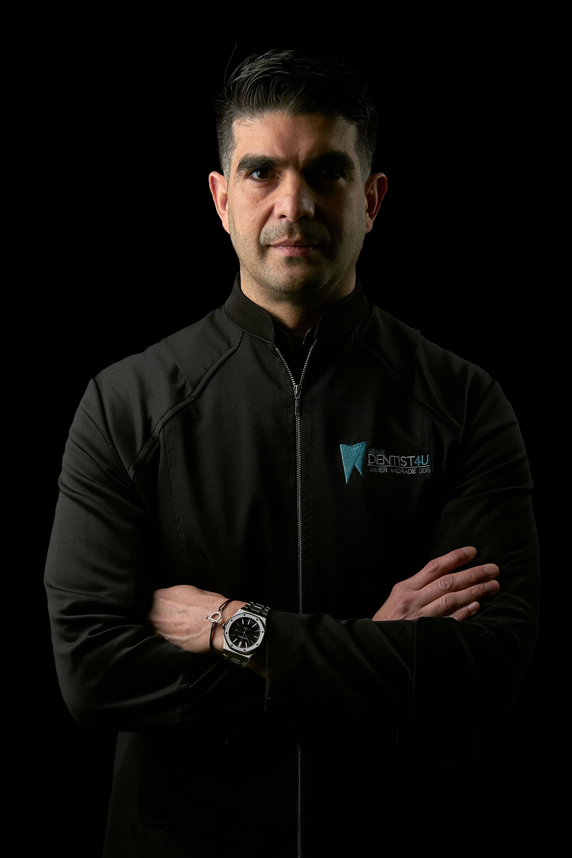 A man in a black jacket is standing with his arms crossed in front of a black background.