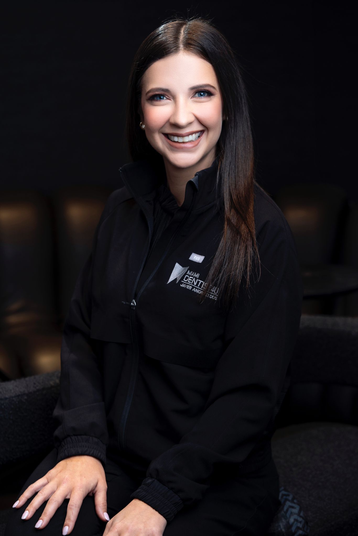 A woman is sitting in a chair wearing a black jacket and smiling.