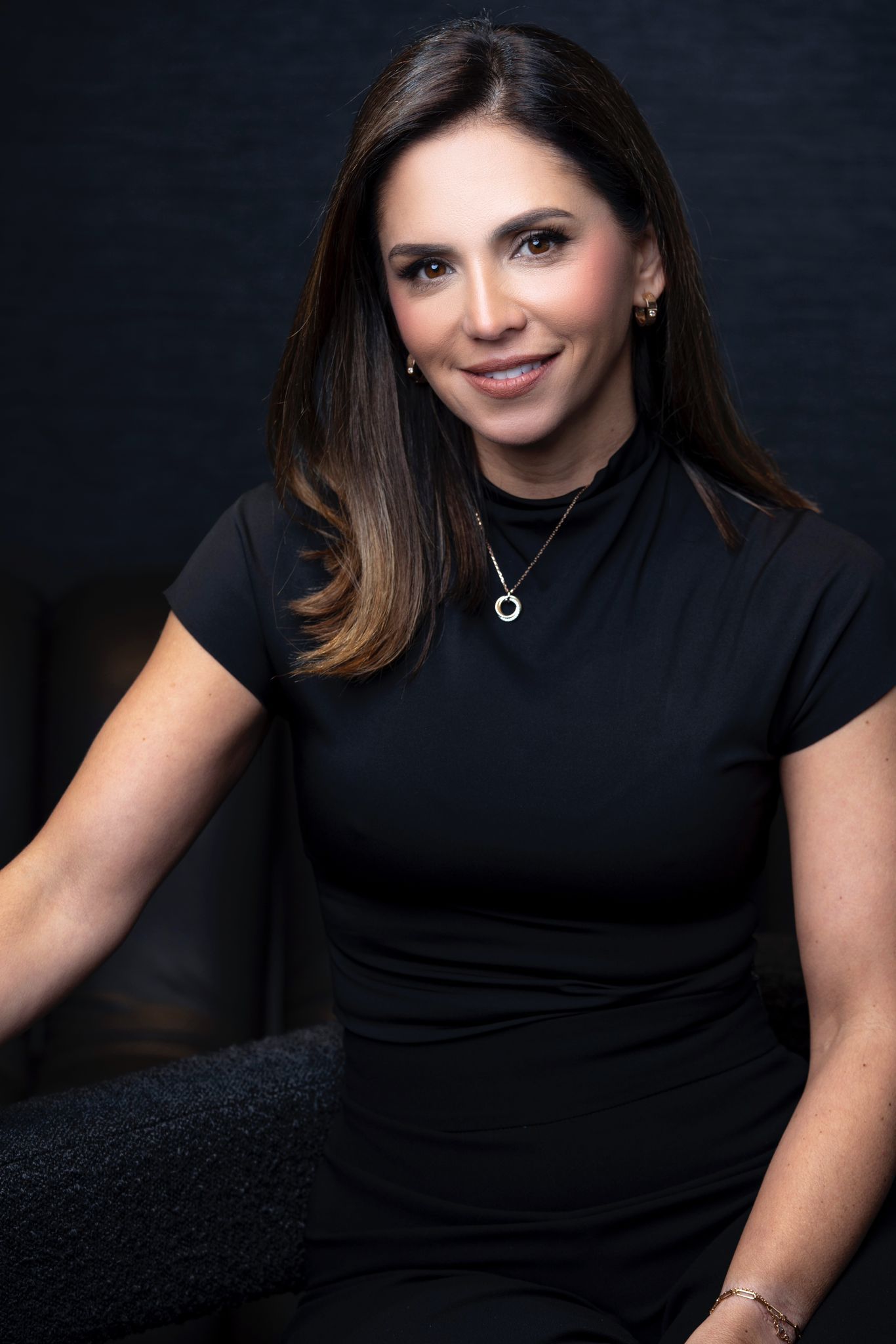 A woman in a black dress is sitting on a chair and smiling.