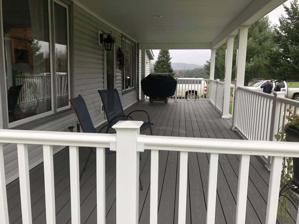 Covered front porch with gray decking, white railings, black chairs, and a grill overlooking a yard.
