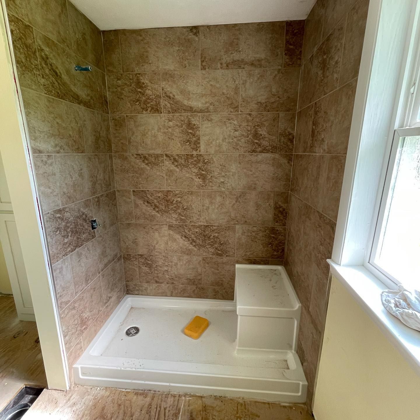 Small tiled bathroom shower stall with white base, built-in shelf, and a window on the right.