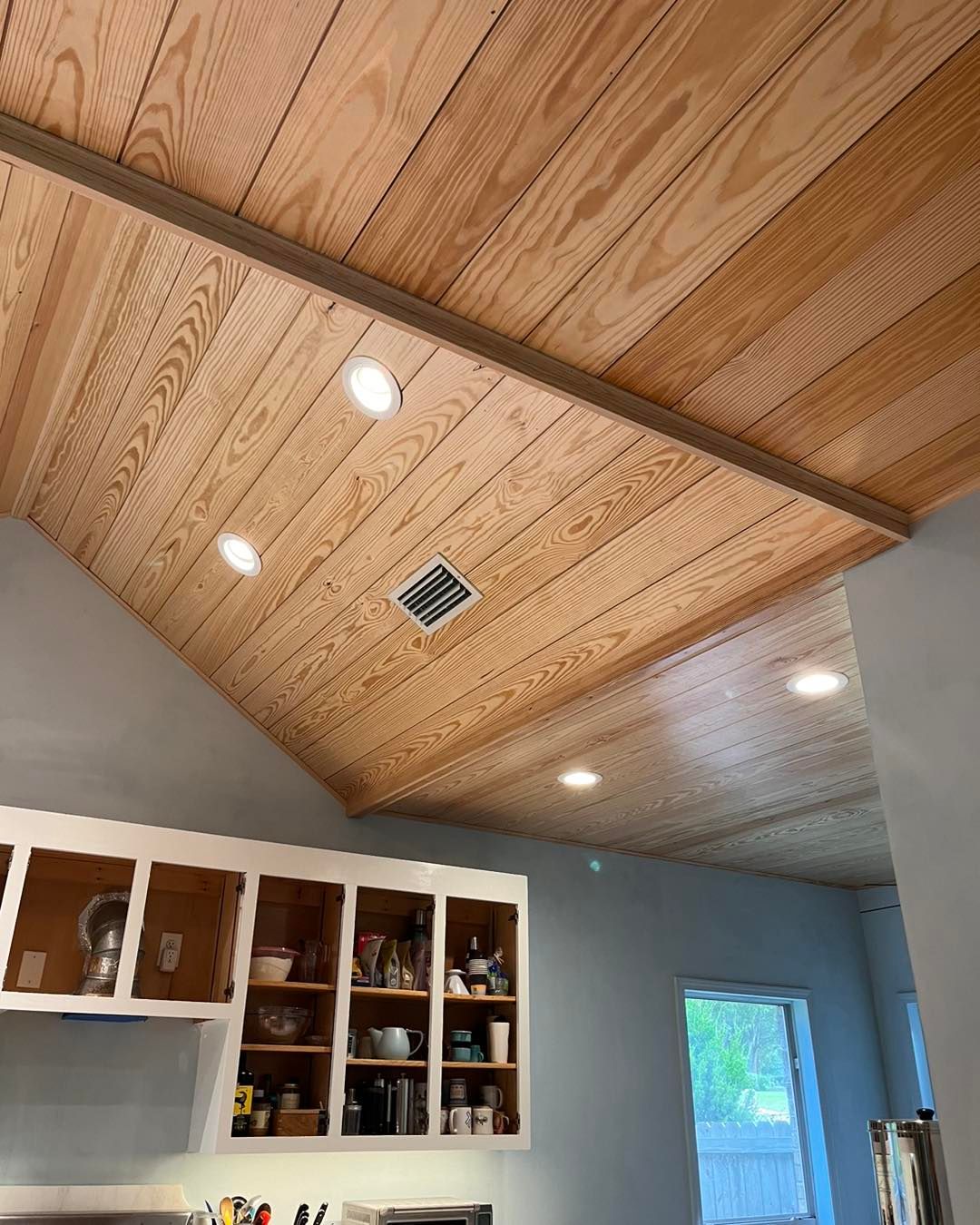 Vaulted wood-paneled ceiling with recessed lights over a kitchen and gray wall.