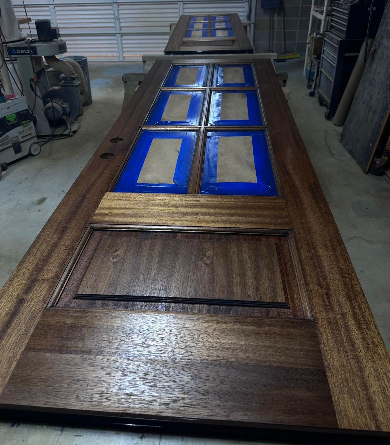 Wooden door with six blue-taped window panes in a workshop, shown lengthwise on a workbench.