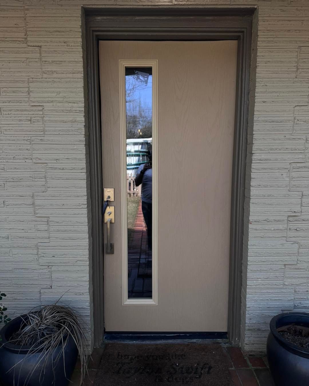 Beige front door with narrow vertical glass panel between two black planters.