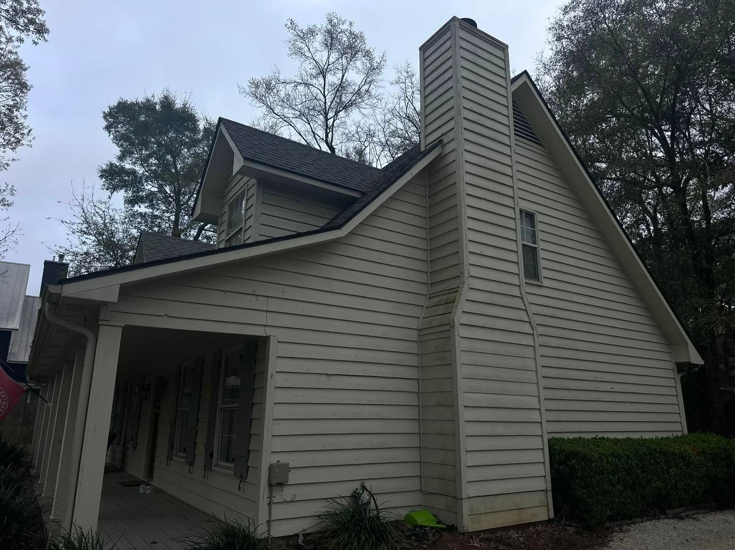 Small beige house with a chimney and covered porch, viewed from the side amid trees.