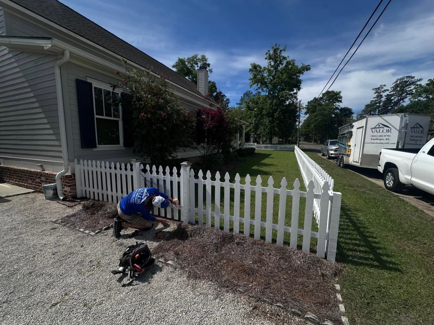 Worker fixing a white picket fence beside a house, with a truck parked on the street.