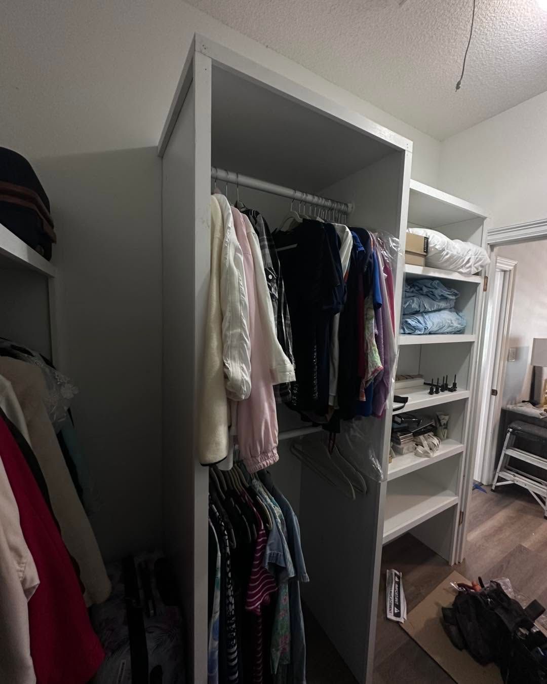 Open white closet with hanging clothes and folded items in a bedroom, shoes on the floor nearby