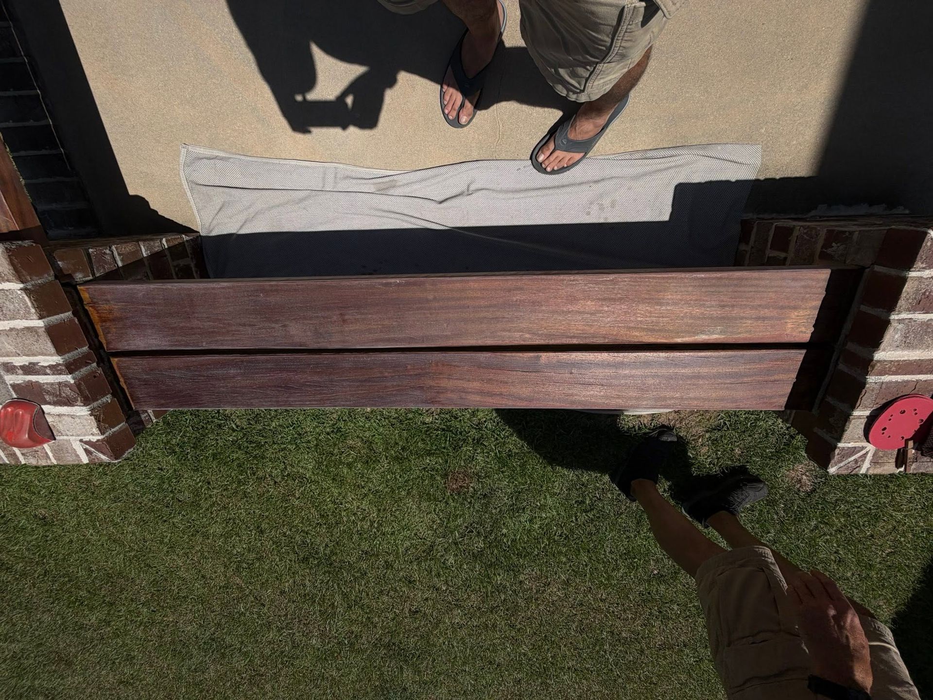 Overhead view of a wooden fence beside a concrete patio and green lawn, with two people’s feet visible.