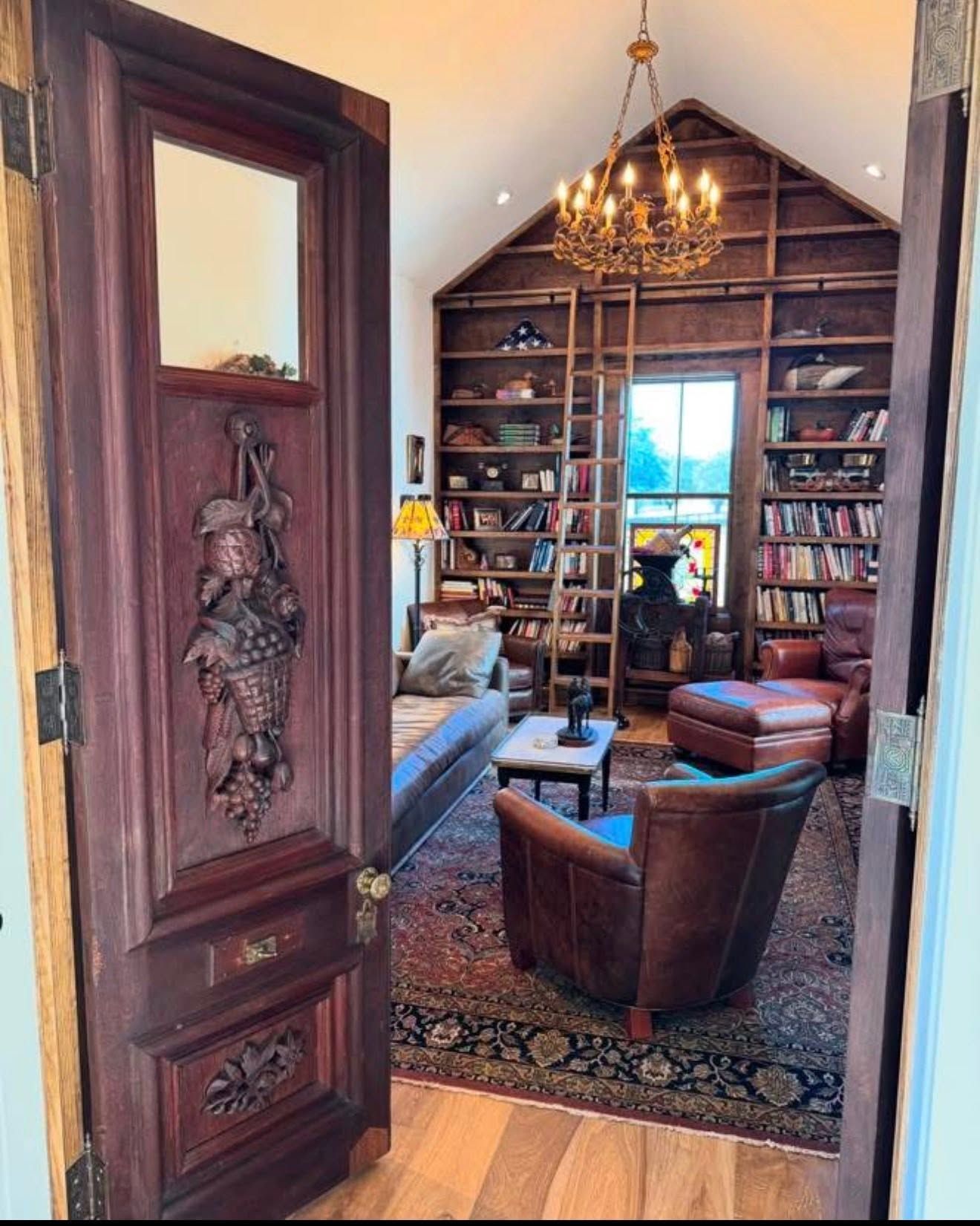 Cozy library with a high bookshelf wall, chandelier, leather chairs, and a person seated near the window.