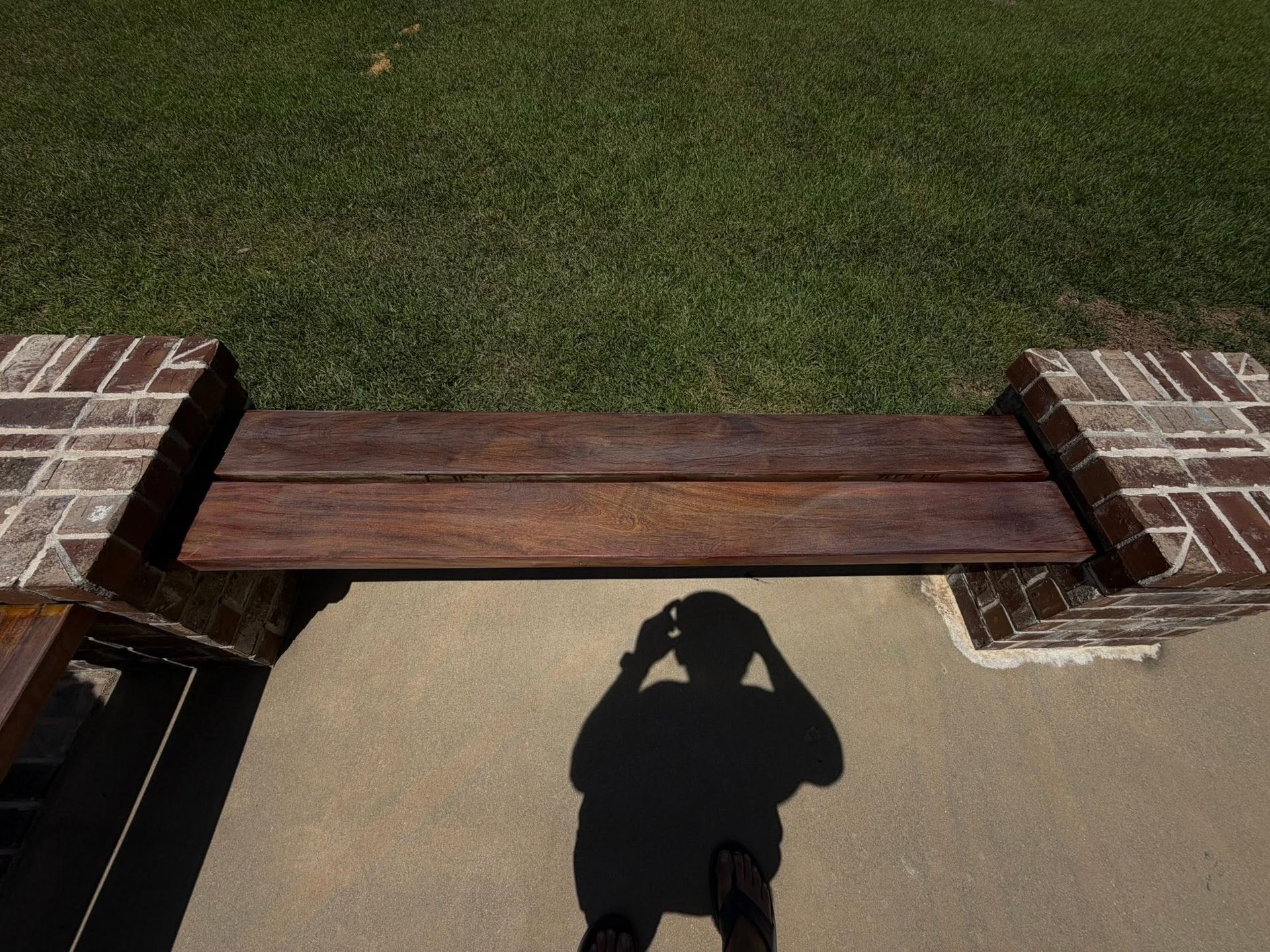 Wooden bench spanning two brick planters beside a sidewalk, with a person’s shadow on the concrete.