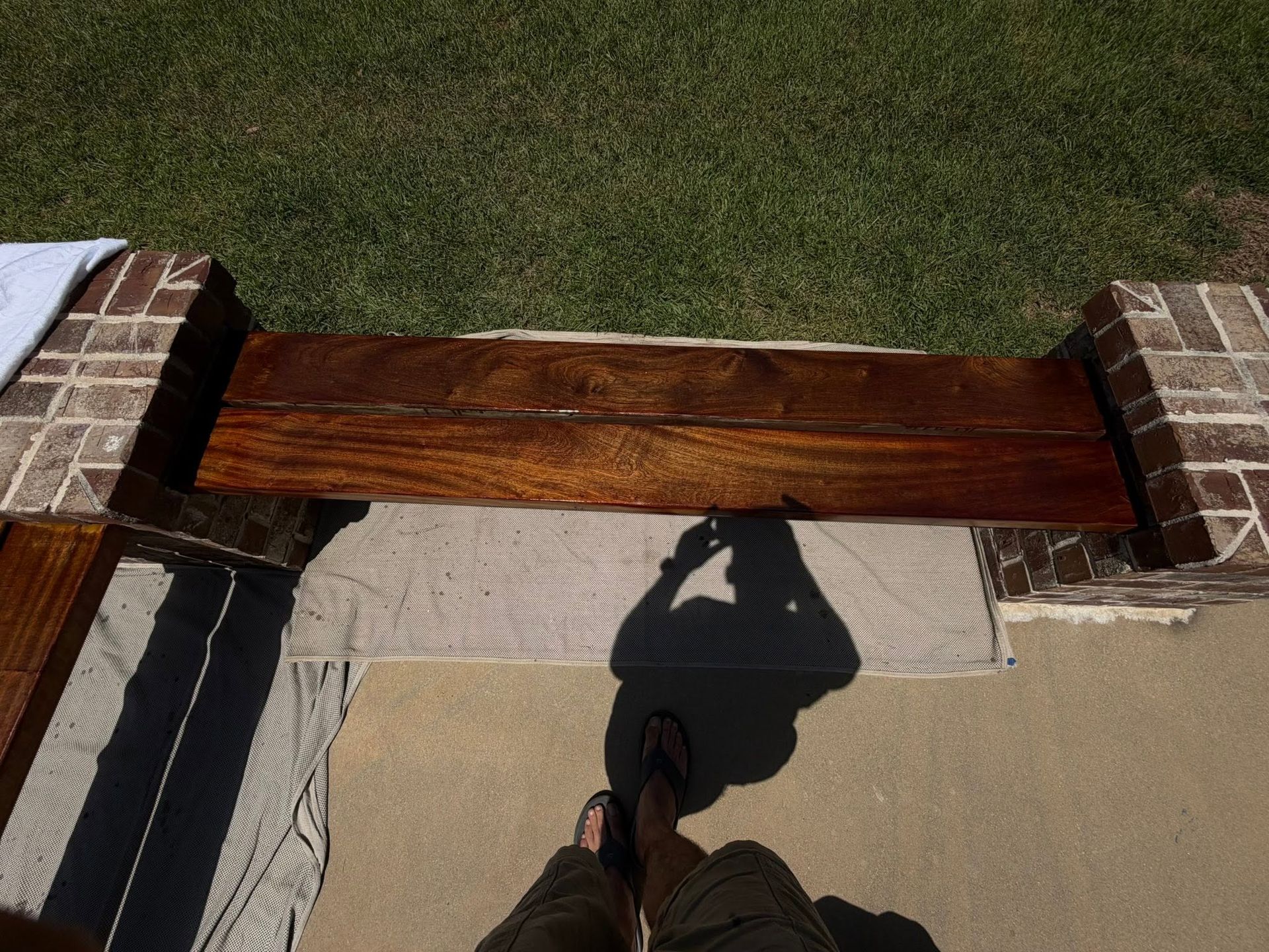 View from a porch step toward a brick-and-wood railing, beige mat, grass, and a person’s shadow on the ground
