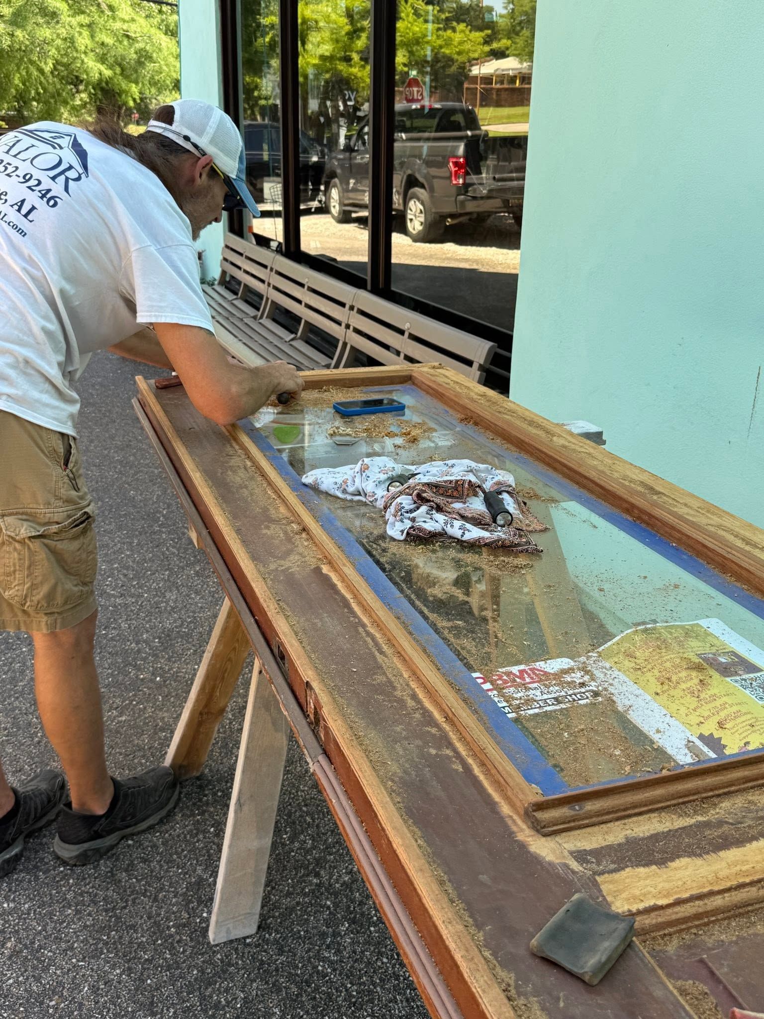 Man leaning over a framed artwork on a table outdoors, inspecting or repairing it.