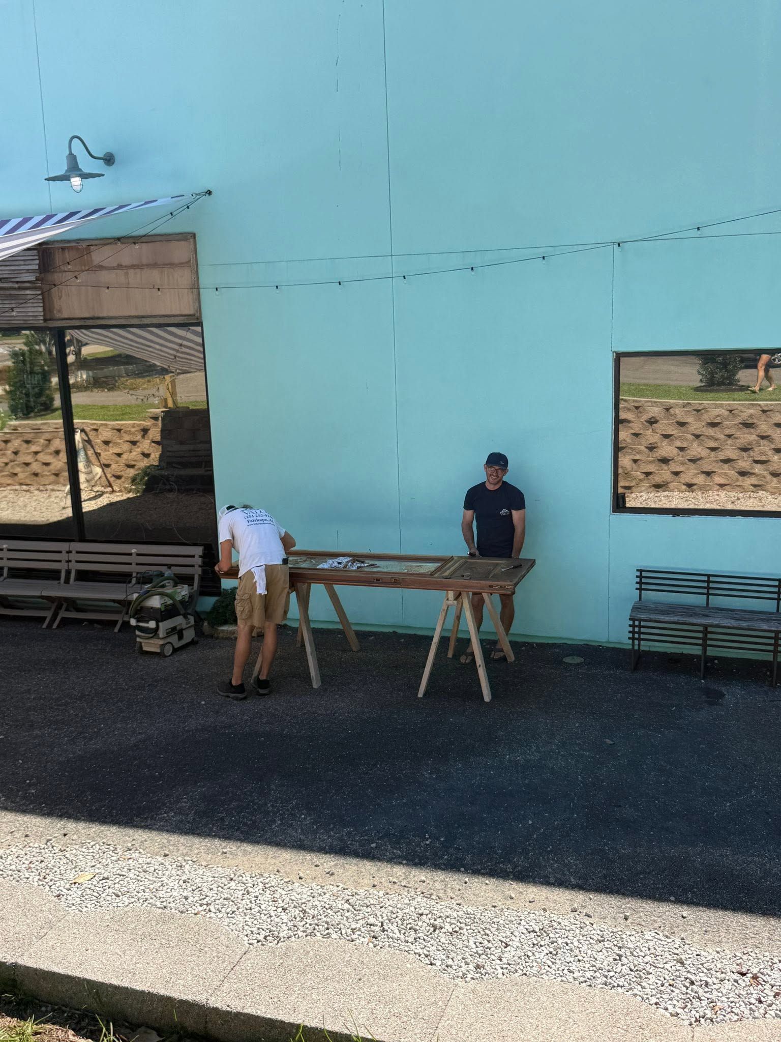 Two people standing by a long wooden table outside a light blue building with open windows
