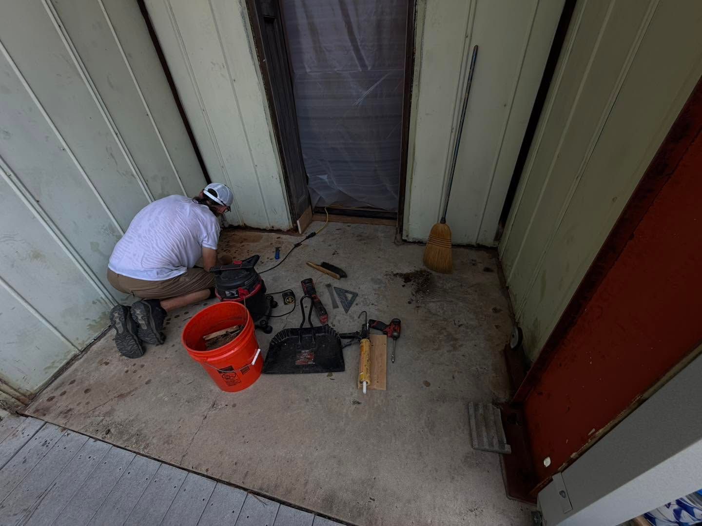 Worker kneeling on porch, using tools beside an orange bucket and open doorway