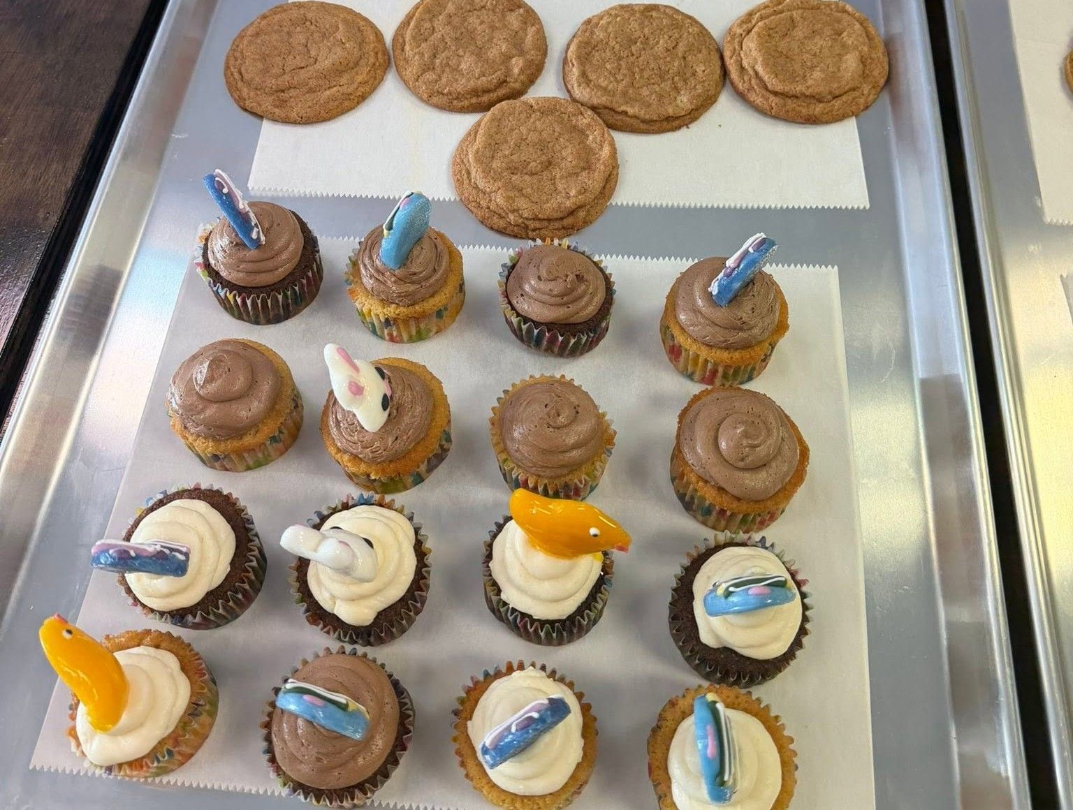 Cupcake tray with frosted mini cakes topped with blue, orange, and white decorations, plus cookies at the top