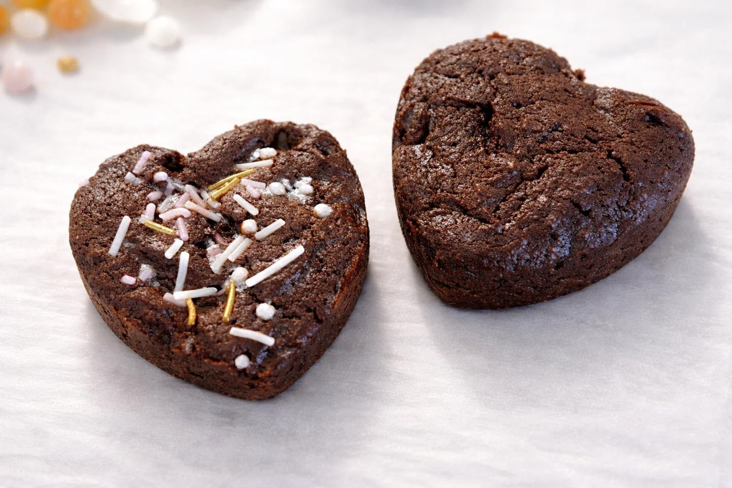 Two heart-shaped chocolate brownies, one topped with sprinkles, on white parchment paper