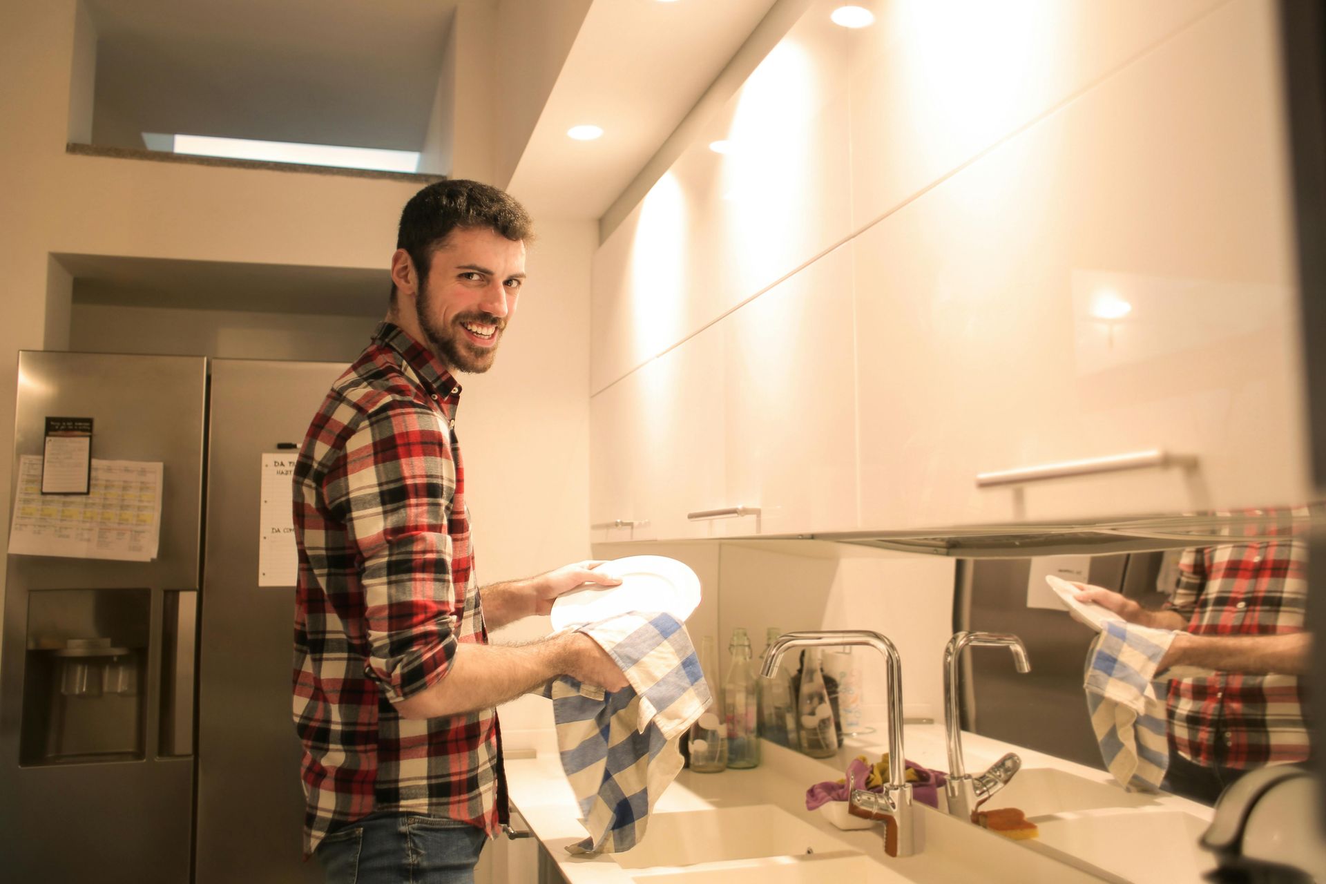 Man drying dishes in a modern kitchen. He smiles, wearing a plaid shirt and jeans.